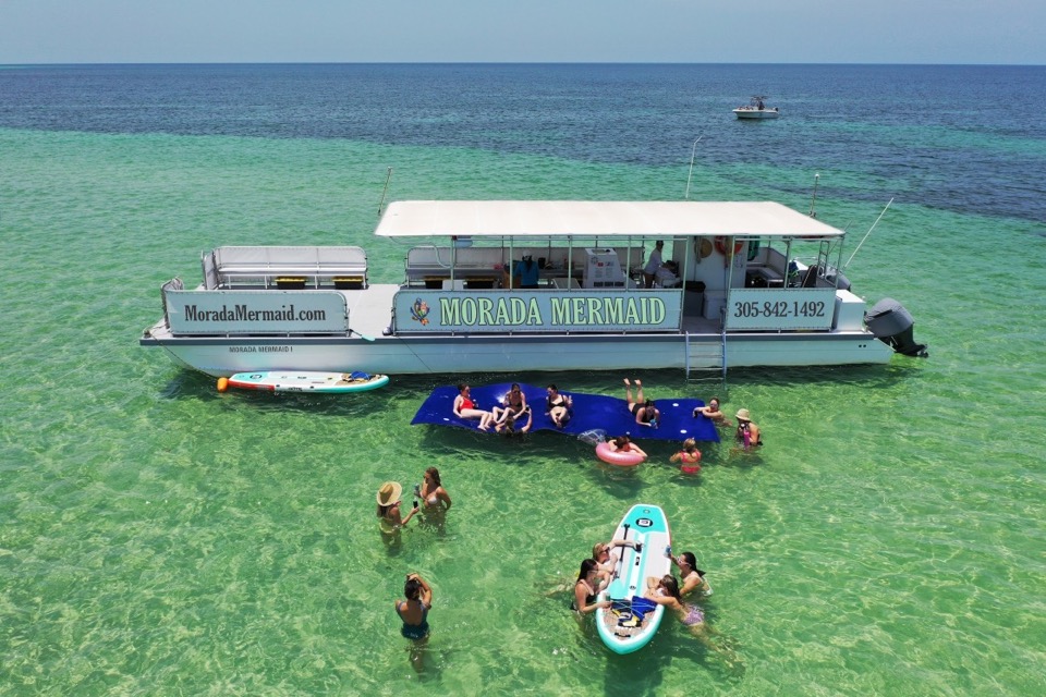 An aerial view of the Morada Mermaid catamaran over turquoise water with guests on floating mats and paddleboards