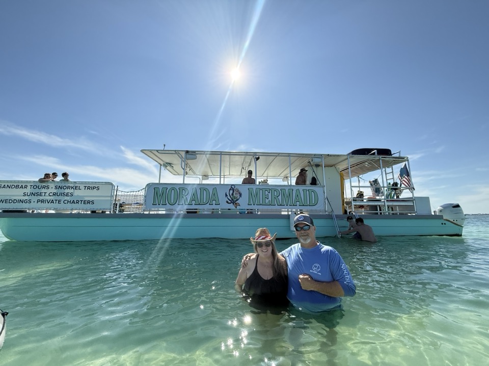 A couple standing in the water in front of the Morada Mermaid with its banner fully readable