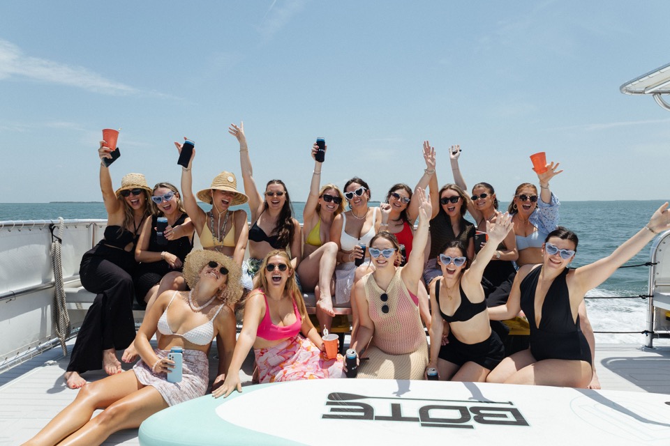 A lively group of women cheering with drinks on the bow under a bright blue sky