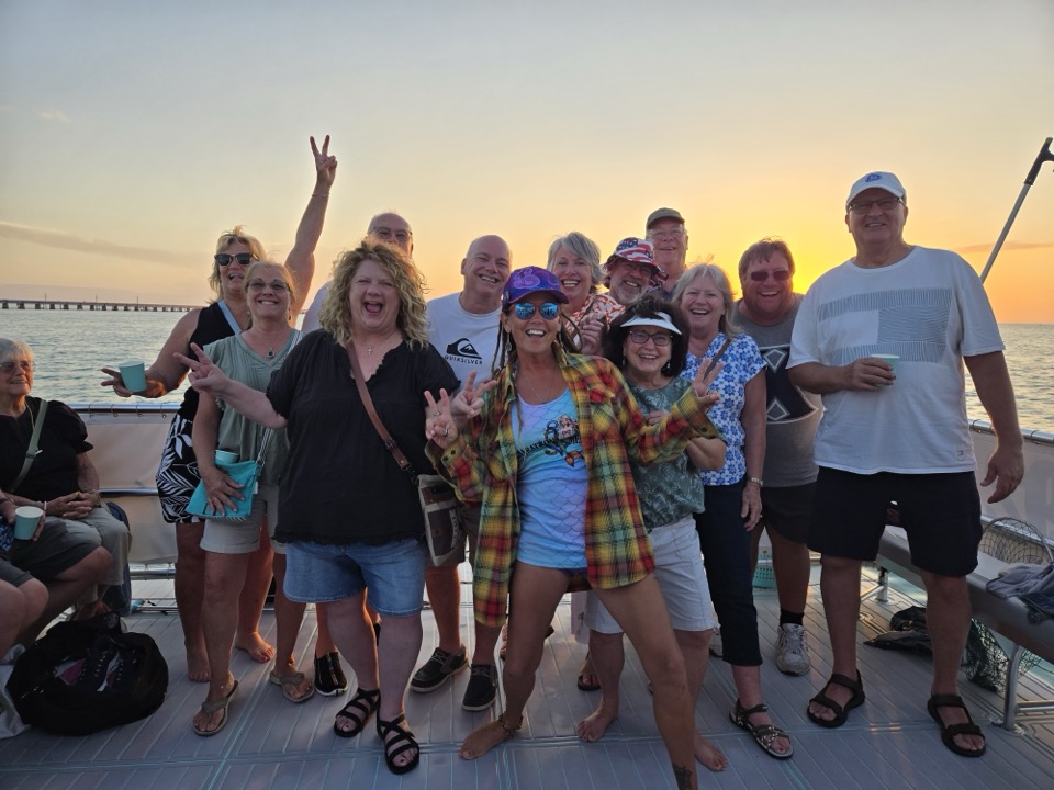 A group of adult guests cheering on deck at sunset with the bridge in the distance