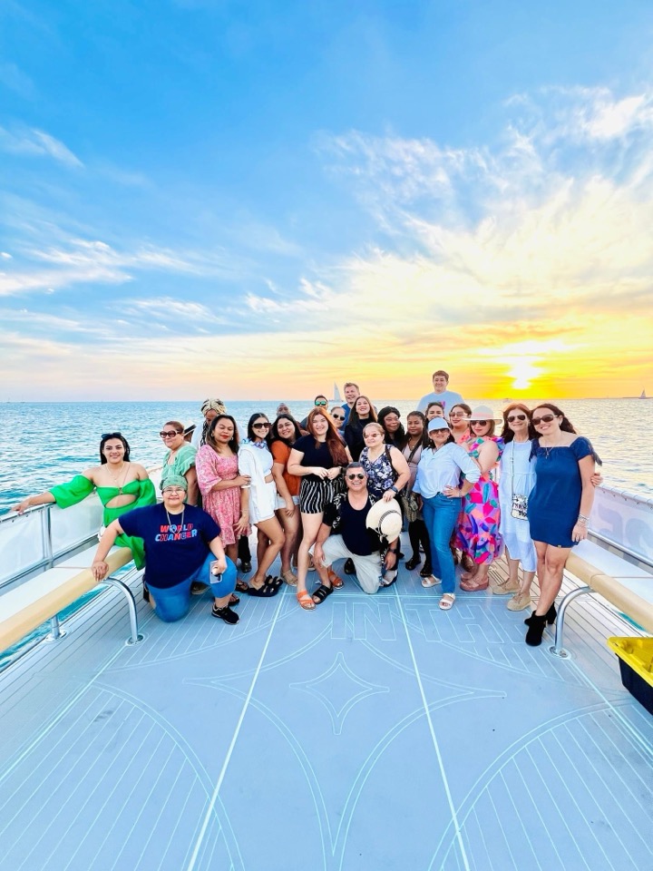 A vibrant large group posing on the catamaran bow at sunset under a blue sky
