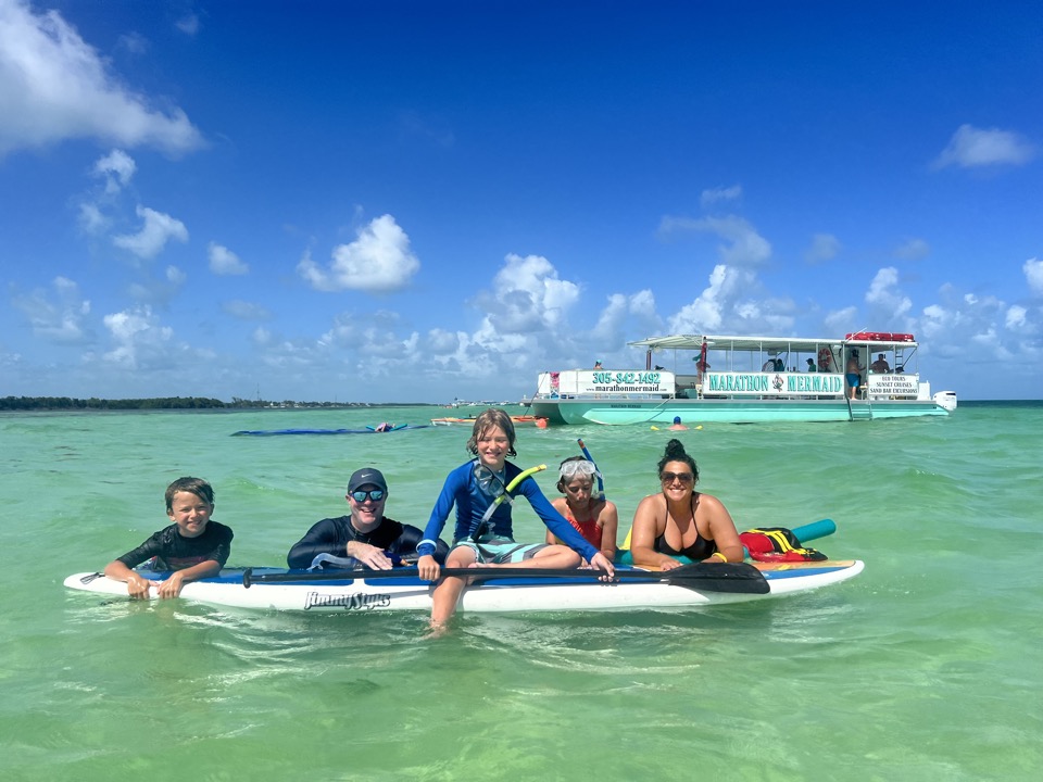 A family of five on a paddleboard with the Marathon Mermaid catamaran anchored behind them in turquoise water