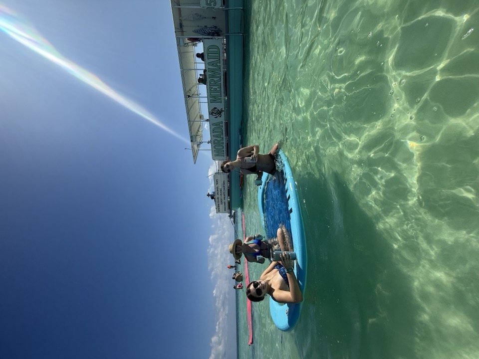 Guests on a paddleboard beside the Morada Mermaid hull in crystal-clear water lit by a sunbeam