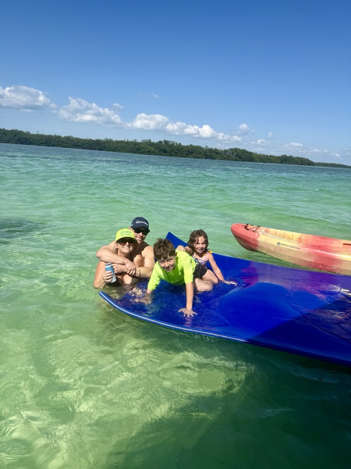 A family smiling on a blue floating water mat with a kayak in gorgeous green sandbar water