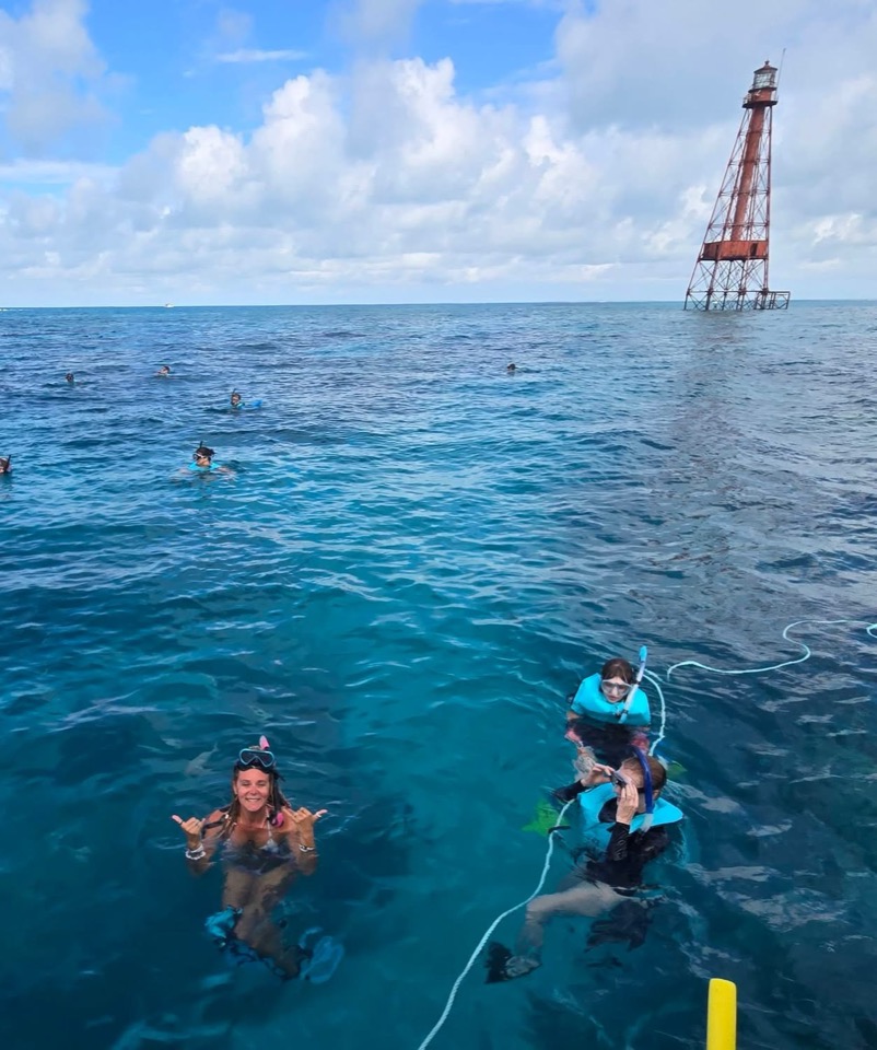 Snorkelers on a line at a reef lighthouse in deep blue water, one throwing a shaka