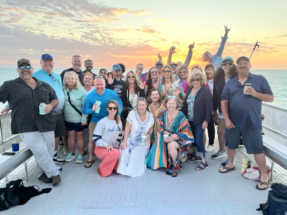 A large group waving with drinks in hand at golden sunset on the boat deck