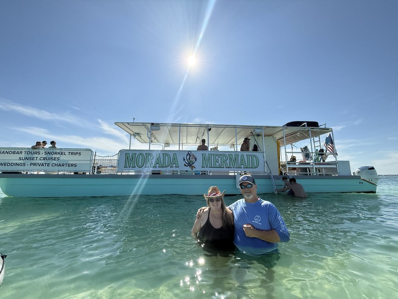 A couple standing in the water in front of the Morada Mermaid with its banner fully readable