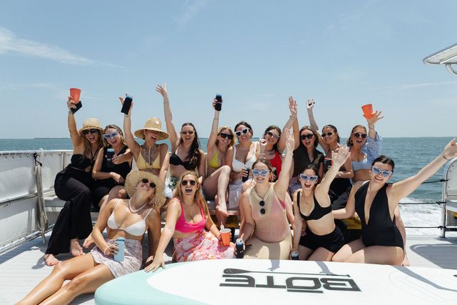 A lively group of women cheering with drinks on the bow under a bright blue sky