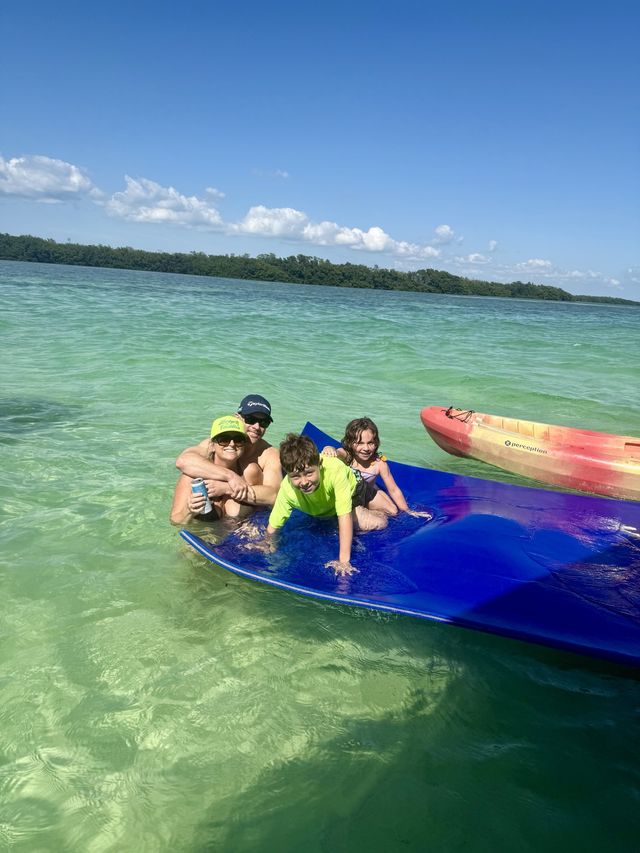 A family smiling on a blue floating water mat with a kayak in gorgeous green sandbar water