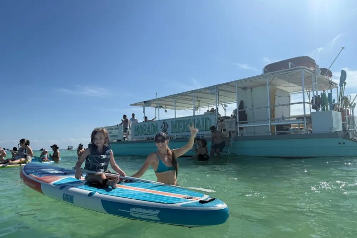 Woman and child on paddleboard near boat named Morada Mermaid in clear, sunny waters.